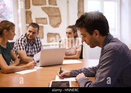 Jeune homme professionnel contrôle ses notes sur sa tablette pendant que le reste de l'équipe sont à la recherche à un ordinateur portable à l'arrière-plan Banque D'Images