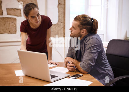 Deux jeunes gens d'affaires à l'un l'autre de façon réfléchie dans l'ordinateur portable qu'ils partagent sur le bureau en face d'eux Banque D'Images