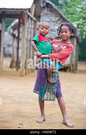 Smiling portrait d'une petite fille malgache tenant son frère dans ses bras à l'avant de cabane en bois pris dans les régions rurales de la communauté de pêcheur (village) Banque D'Images