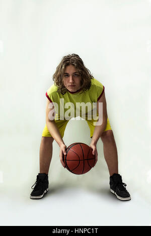 Jeune garçon avec de longs cheveux blonds portant un maillot vert dans une position accroupie tenant une basket-ball Banque D'Images