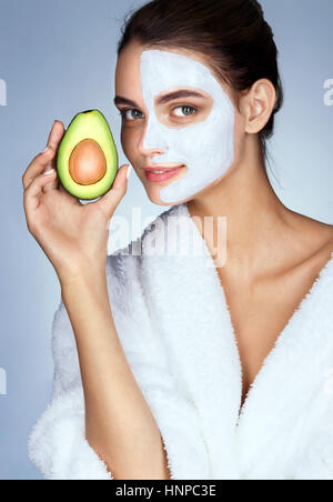 Jolie Jeune femme tenant une demi-avocat en main. Photo de femme avec masque facial hydratant. Beauté et Soins De La Peau concept Banque D'Images