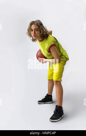 Jeune garçon avec de longs cheveux blonds portant un maillot vert dans une position accroupie tenant une basket-ball Banque D'Images