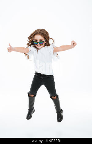 Toute la longueur de smiling little girl à lunettes de saut et showing Thumbs up over white background Banque D'Images