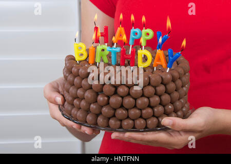 Young Girl holding anniversaire chocolat gâteau décoré de bonbons boules de malt en face d'elle avec joyeux anniversaire bougies. Facile et rapide gâteau fait maison Banque D'Images