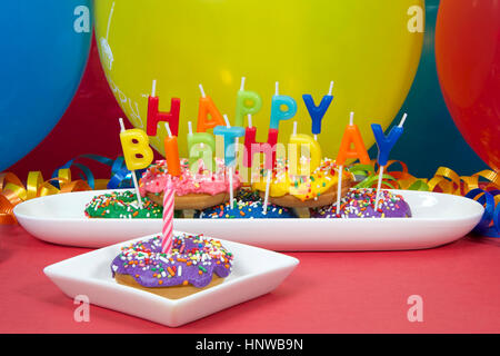 Beignet miniature gâteau pour première fête d'anniversaire. Multicolor donuts sur plaque blanche avec Joyeux Anniversaire bougies. Un beigne givré mauve cupcake on plate Banque D'Images