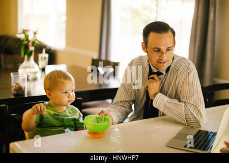 Le père et le jeune fils à table de cuisine, son eating breakfast, père de mettre sur collier, looking at laptop Banque D'Images