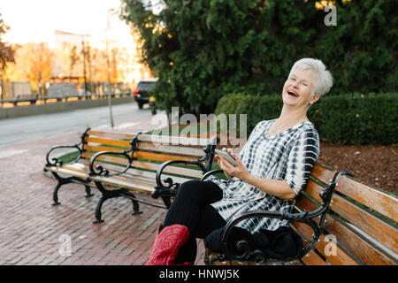 Portrait of senior woman, à l'extérieur, assis sur un banc, holding smartphone, rire Banque D'Images