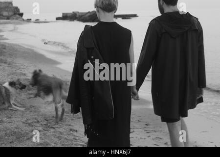 Jeune couple en train de marcher le long de la plage main dans la main, deux chiens en avance, vue arrière Banque D'Images