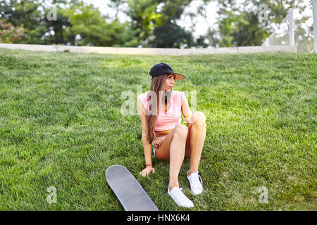 Jeune femme assise sur la planche à roulettes, herbeux à côté d'elle Banque D'Images
