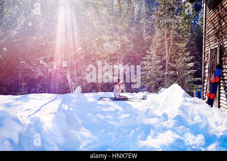 Woman practicing yoga, à genoux sur un tapis de yoga par log cabin en neige, Autriche Banque D'Images