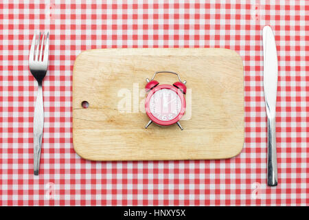 Le temps de manger Concept : 12 o'clock Horloge rouge sur un bloc en bois, couteau et fourchette sur rouge checker table cloth Banque D'Images