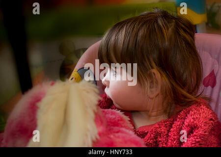 Petite fille blonde assise sur une balançoire, portant un chandail de fourrure rose et à la recherche sur le côté tout en tenant un ours en peluche. Banque D'Images