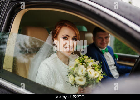 Mariée aux cheveux rouge sourit siiting avec mariés à voiture et à la recherche à partir de la fenêtre, à la pluie. Banque D'Images