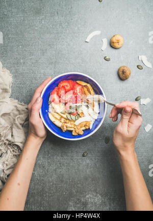 Le Yogourt, Céréales, graines, fruits secs et frais et le miel dans un bol en céramique bleu femme' s les mains sur fond gris, vue de dessus, copiez l'espace. Manger propre Banque D'Images