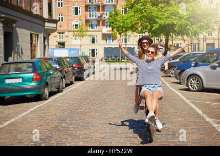 Jeune femme pétillante équitation sur le guidon d'un vélo d'amis comme le billet le long d'une rue pavée de la ville de rire à l'appareil photo Banque D'Images