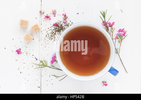 Une tasse bleue avec tisane sur un fond de bois blanc. La tasse est entouré de fleurs roses, cubes de sucre de canne et de feuilles de thé. Banque D'Images