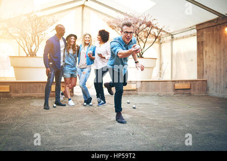 Jeune homme séduisant en acier Lunettes jeter Boule de petanque, ses amis smiling standng derrière lui Banque D'Images