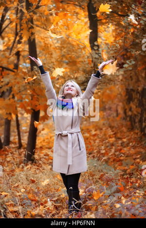 Laughing girl avec de longs cheveux blonds jette feuilles à l'automne parc. Femme heureuse de jouer avec les feuilles d'automne en forêt. Concept loisirs insouciant bonheur Banque D'Images