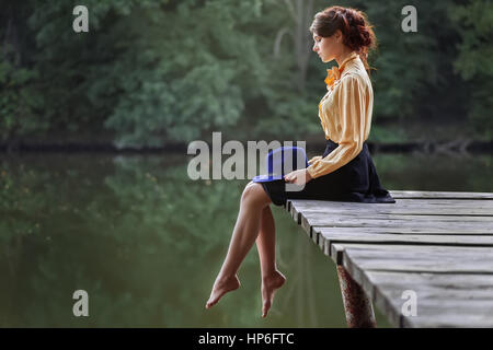 Vue latérale du dreaming girl sitting on jetty forêt et rivière sur l'arrière-plan. Belle femme assise au bord du lac. Sad girl déprimé assis sur pont. Banque D'Images