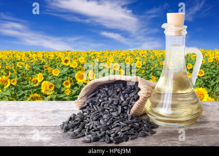 L'huile de tournesol dans le flacon en verre et les graines sur table en bois avec champ de fleurs sur l'arrière-plan. Champ de tournesol avec ciel bleu Banque D'Images