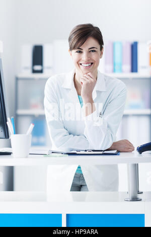 Attractive young woman leaning on la clinique/24 with hand on chin, il est smiling at camera, le personnel médical et les soins de concept Banque D'Images