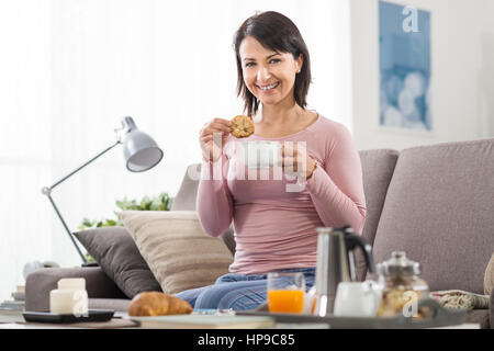 Femme détendue sereine de prendre le petit déjeuner le matin, elle est assise sur le canapé et trempage des cookies dans son café Banque D'Images