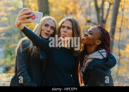 Trois superbes happy woman in autumn park selfies. Jolies filles avec différentes couleurs de peau. Femme faire des grimaces et smiling at camera. Banque D'Images