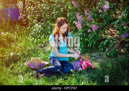 Jeune femme assise dans le parc à l'herbe avec des fleurs de lilas et tulipes Banque D'Images