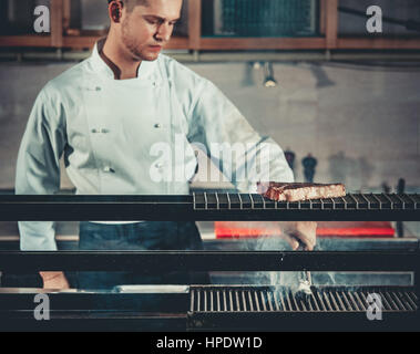 Jeune chef blanc en tablier, debout près du brasier avec charbons. Man cooking steak de boeuf dans l'intérieur de cuisine professionnelle moderne Banque D'Images