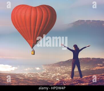 Fille aux mains levé sur le sommet de la montagne à la recherche de big red air balloon forme de coeur battant en bleu ciel. Le jour de la saint Valentin romantique. Sport et loisir Banque D'Images