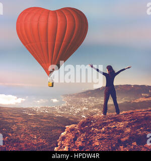 Girl with hands up stand sur le dessus de la montagne à la recherche de big red air balloon forme de coeur battant en bleu ciel. Voyage Romantique le jour de la Saint-Valentin. Sports, t Banque D'Images