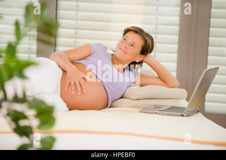 Parution , Hochschwangere modèle Frau liegt am gemuetlich Bett, ordinateur portable steht daneben - pregnant woman with laptop relaxing in bed Banque D'Images