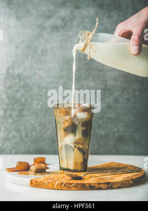 La main de l'homme de verser le lait de la bouteille de verre avec caramel glacé et café congelé des cubes de glace sur le bois d'olive et en servant au tableau gris, dar Banque D'Images