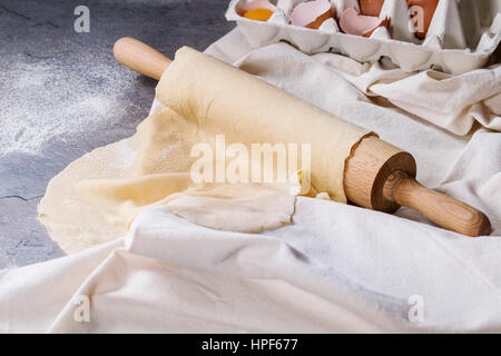 Des pâte roulée pour les pâtes tagliatelle sur bois rouleau à pâtisserie avec le jaune d'œuf et le linge blanc gris foncé dessus textile fond de table. Banque D'Images