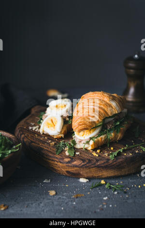 Croissant sandwich au thon avec oeuf mollet, roquette, salade de thon et de concombre sur planche à découper en bois. Vue rapprochée, selective focus Banque D'Images