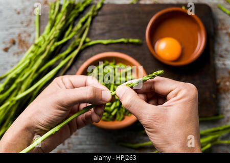 Young man chopping asperges sauvages avec ses mains et des bols en terre cuite avec des morceaux d'asperges et oeuf fissuré, les ingrédients pour préparer un comme Banque D'Images