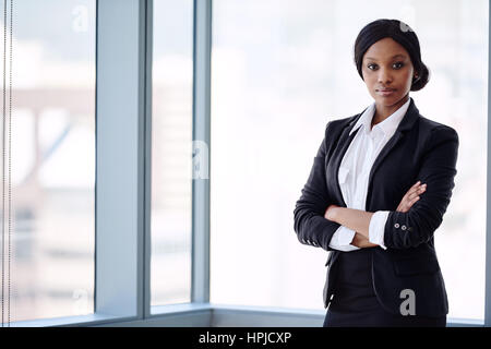 Femme d'affaires africains habillés formellement à la recherche dans l'appareil photo avec les bras croisés en se tenant debout en face de grandes fenêtres en verre avec un paysage urbain Banque D'Images