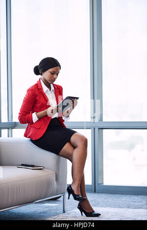 Jeune femme noire assise sur le bord d'un canapé dans un salon d'affaires tout en regardant l'écran de la tablette numérique qu'elle est tenue à Banque D'Images