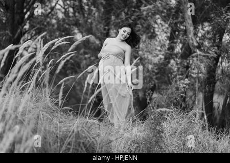 Belle femme enceinte dans une robe turquoise à l'extérieur. Les arbres en arrière-plan. L'herbe haute à l'avant-plan. Photo en noir et blanc. Banque D'Images