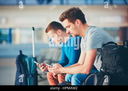 Deux amis voyageant ensemble. Voyageurs avec des téléphones mobiles en attente à l'aéroport, zone de départ de leur vol pour retard. Banque D'Images