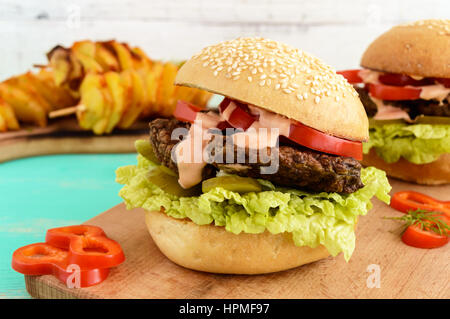 Escalope de foie avec des hamburgers, des tomates, des cornichons, de la laitue, sauce épicée et un pain avec des graines de sésame sur une planche à découper et de tranches de pommes de terre en brochettes. Banque D'Images