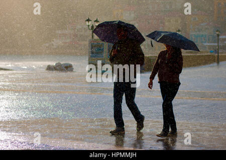 Couple en silhouette marcher le long de la rue pavée par la mer à Parga, Grèce avec des parapluies dans la pluie comme du soleil apparaît Banque D'Images