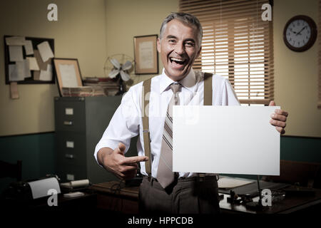 Cheerful businessman showing a blank sign and smiling, 1950 office sur l'arrière-plan. Banque D'Images