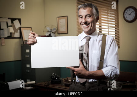 Cheerful businessman showing a blank sign and smiling, 1950 office sur l'arrière-plan. Banque D'Images