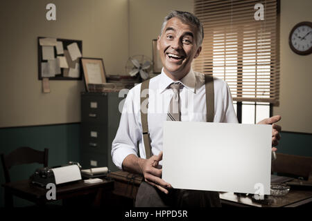 Cheerful businessman showing a blank sign and smiling, 1950 office sur l'arrière-plan. Banque D'Images