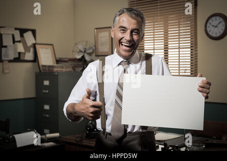 Cheerful businessman showing a blank sign and smiling, 1950 office sur l'arrière-plan. Banque D'Images