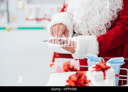 Le Père Noël de faire des achats de Noël au supermarché, il est utiliser des applications sur une tablette numérique et en poussant un chariot rempli de boîtes cadeaux Banque D'Images