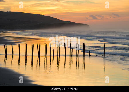 Dunedin, Otago, Nouvelle-Zélande. Vue sur St Clair Beach au lever du soleil, célèbre rangée de pieux en bois reflète dans l'eau. Banque D'Images