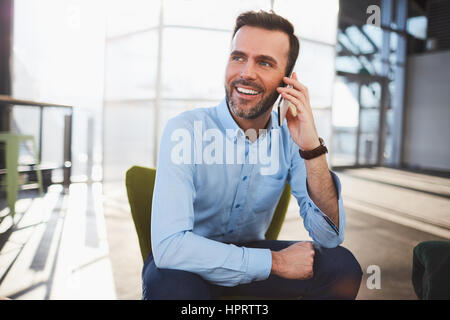 Jeune homme barbu à l'aide d'un téléphone mobile comme il est assis sur une chaise dans un restaurant dans un grand vestibule spacieux à la voiture avec un sourire alors qu'il s'entretient Banque D'Images