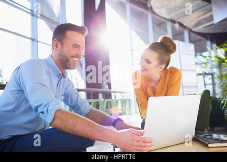 Young smiling couple partageant un ordinateur portable comme ils vous détendre autour d'un café dans un grand hall lumineux et spacieux ou vestibule avec sun flare Banque D'Images